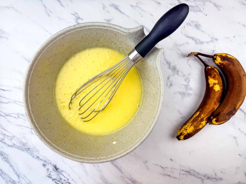 A mixing bowl with a whisk and a yellow batter next to two ripe, spotted bananas on a marble surface.