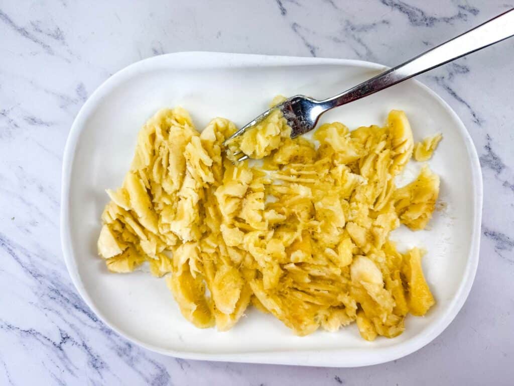 A fork is mashing bananas on a white oval plate, placed on a marble surface.
