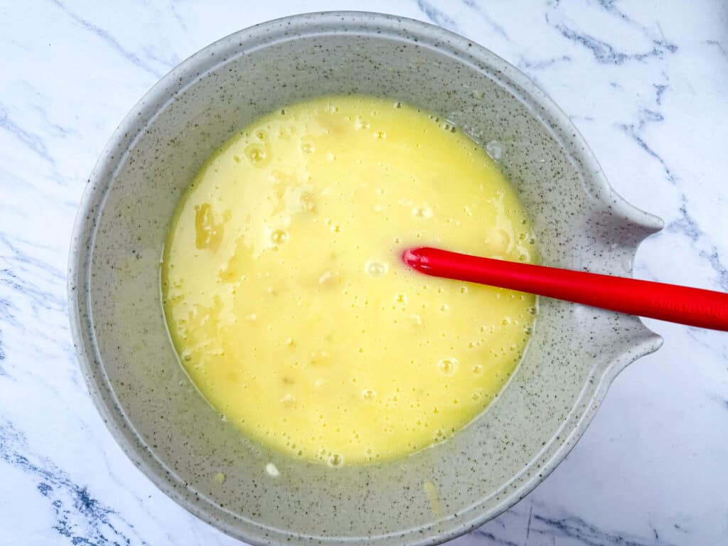 A speckled mixing bowl filled with a yellow liquid mixture and a red spatula on a white marble surface.