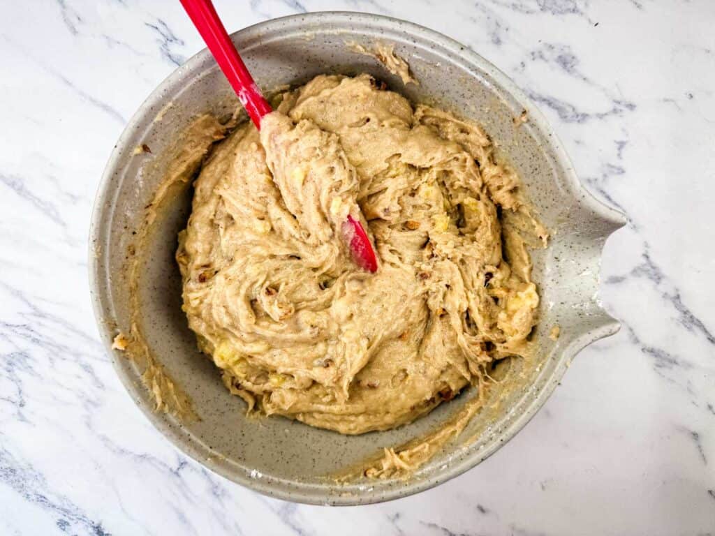A mixing bowl filled with thick banana bread batter, being stirred with a red spatula, sits on a white marble surface.
