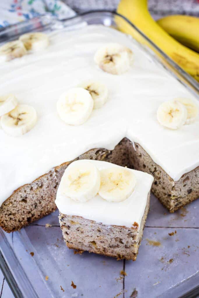 A glass baking dish with banana cake topped with white frosting and banana slices. One piece is cut and placed in front, showing the texture inside. Whole bananas are in the background.