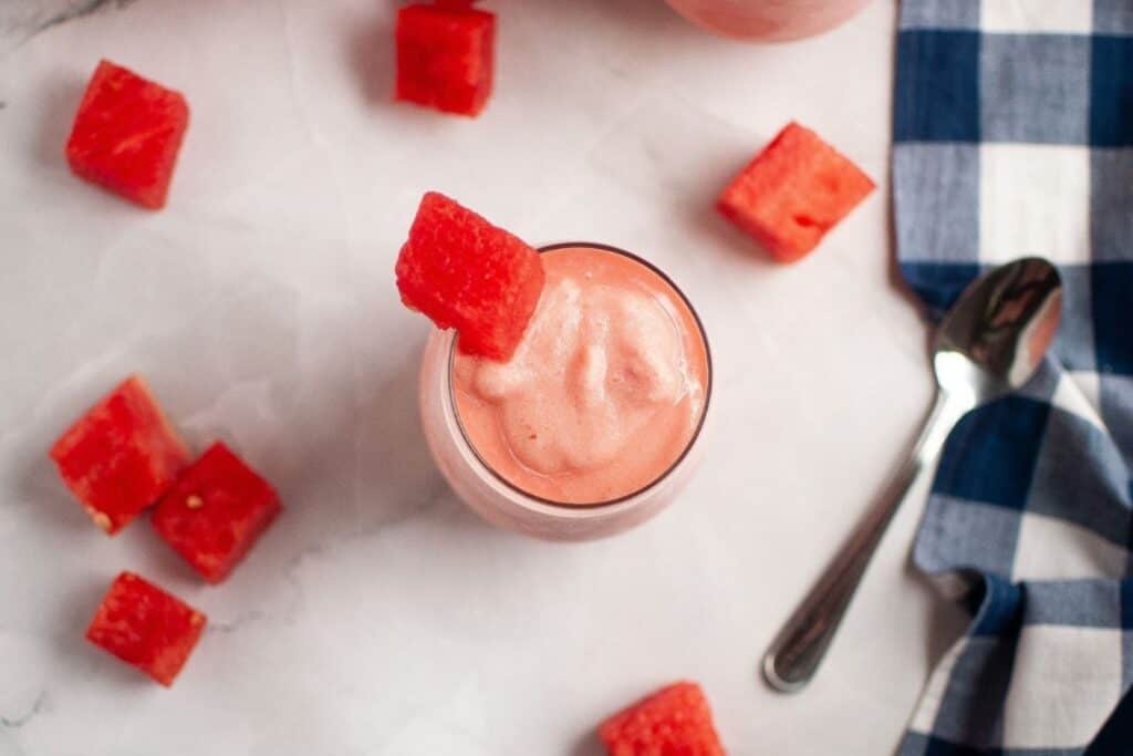 A glass of pink watermelon smoothie topped with a watermelon cube, surrounded by additional watermelon cubes, a spoon, and a blue checkered napkin on a white surface.
