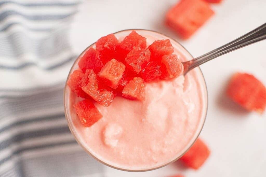 A spoonful of diced watermelon on top of a glass filled with pink frozen dessert, with more watermelon cubes scattered nearby.