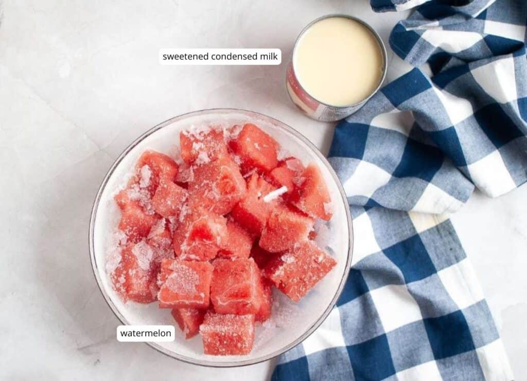 A bowl of frozen watermelon cubes is next to an open can of sweetened condensed milk and a blue-and-white checkered cloth on a white surface.