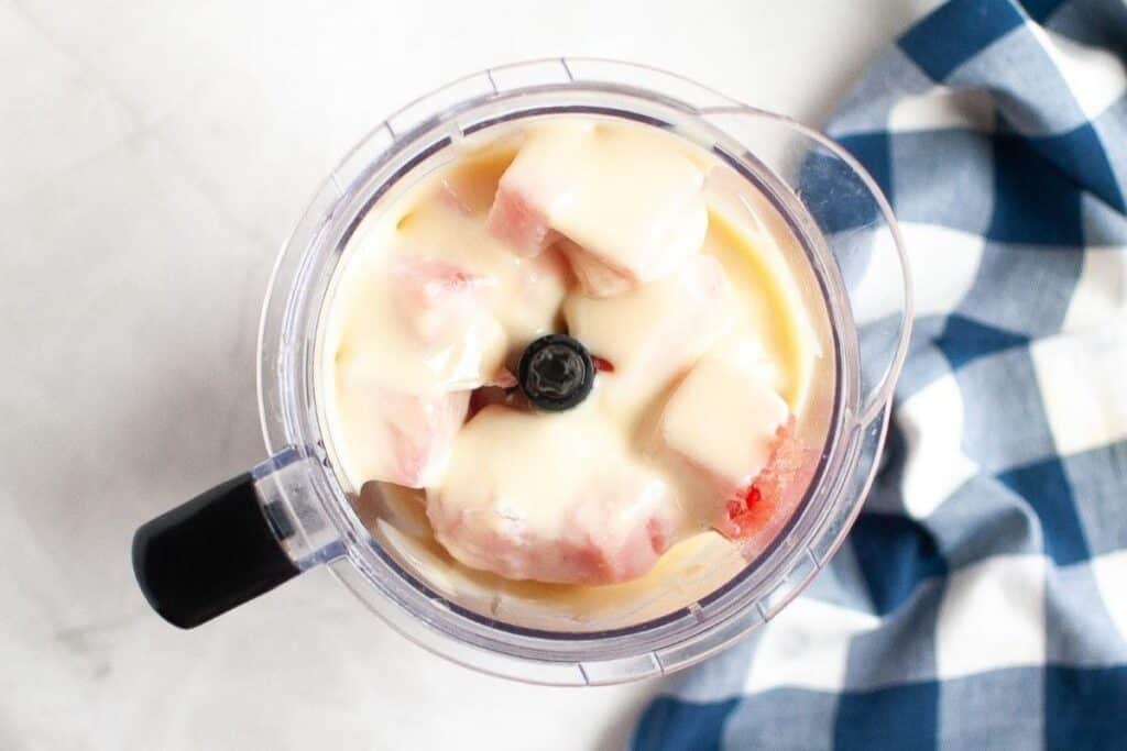 A blender filled with chopped fruit and creamy liquid next to a blue and white checkered cloth on a white surface.