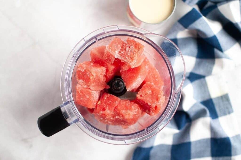 A blender filled with frozen watermelon cubes sits on a light surface next to a blue and white checkered cloth and an open can.