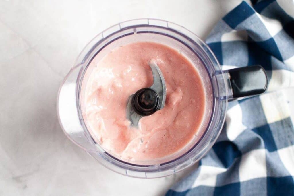 A food processor containing a creamy pink mixture, positioned next to a blue and white checkered cloth on a light surface.