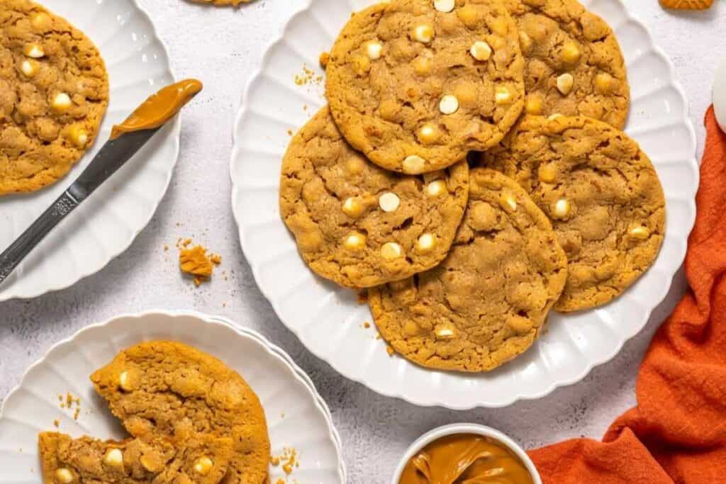 A plate of large cookies with white chocolate chips, a partially eaten cookie on a smaller plate, a knife, and a small bowl of peanut butter on a white surface.