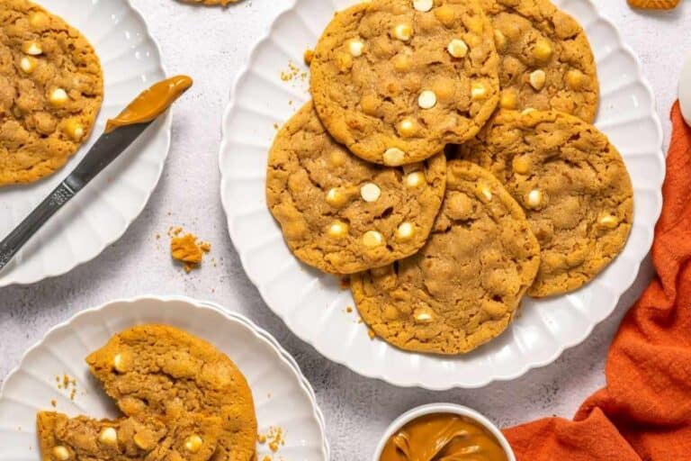 A plate of large cookies with white chocolate chips, a partially eaten cookie on a smaller plate, a knife, and a small bowl of peanut butter on a white surface.