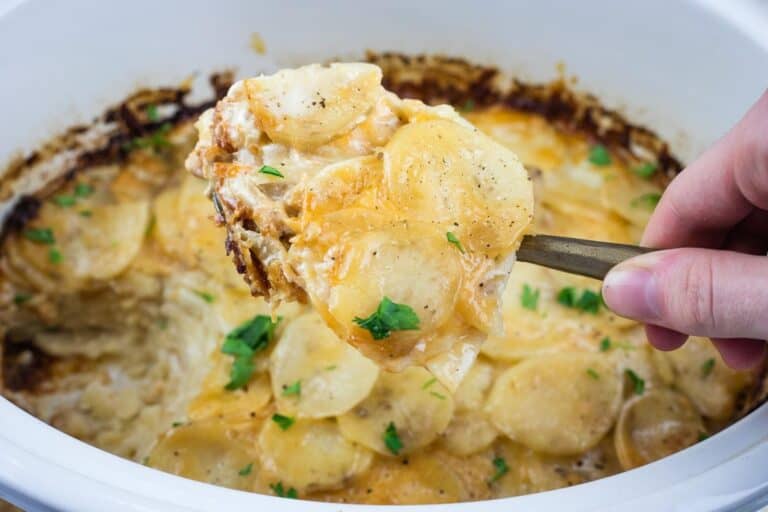 A hand holding a serving spoon with cheesy scalloped potatoes above a casserole dish filled with more scalloped potatoes, garnished with parsley.