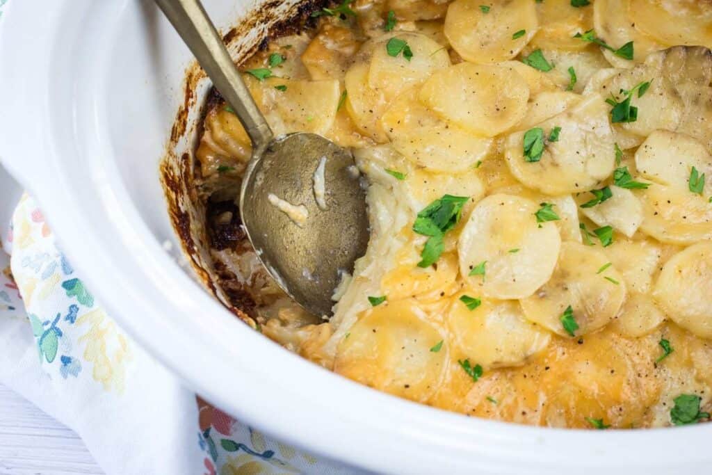 A serving spoon rests in a slow cooker filled with cheesy scalloped potatoes, garnished with chopped parsley.