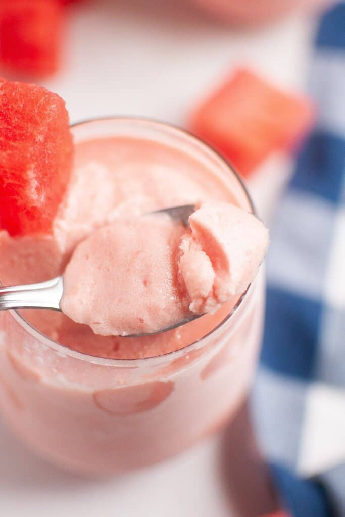 A spoonful of pink watermelon sorbet is held above a glass jar, with a watermelon slice on the rim and a blue checkered cloth in the background.