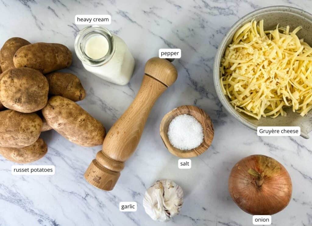 Ingredients for a recipe arranged on a marble surface: russet potatoes, garlic, onion, Gruy&egrave;re cheese, heavy cream, salt, pepper.
