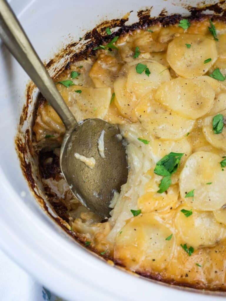 A close-up of cheesy scalloped potatoes in a white dish, with a serving spoon and chopped parsley sprinkled on top.