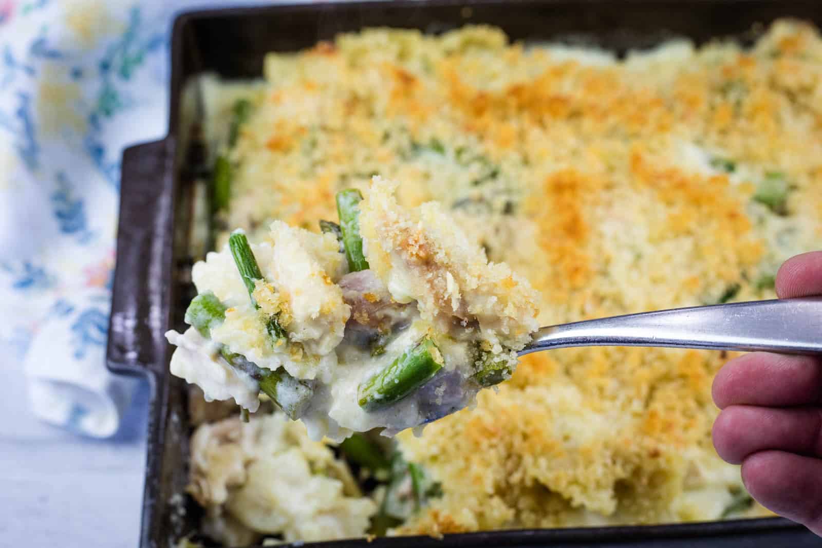 A hand holds a spoonful of Gruy&egrave;re Chicken Asparagus Pasta casserole topped with breadcrumbs over a baking dish filled with the same casserole.
