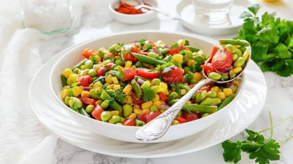 A white bowl filled with a colorful mixed vegetable salad, including corn, tomatoes, green beans, and edamame, with a spoon resting inside.