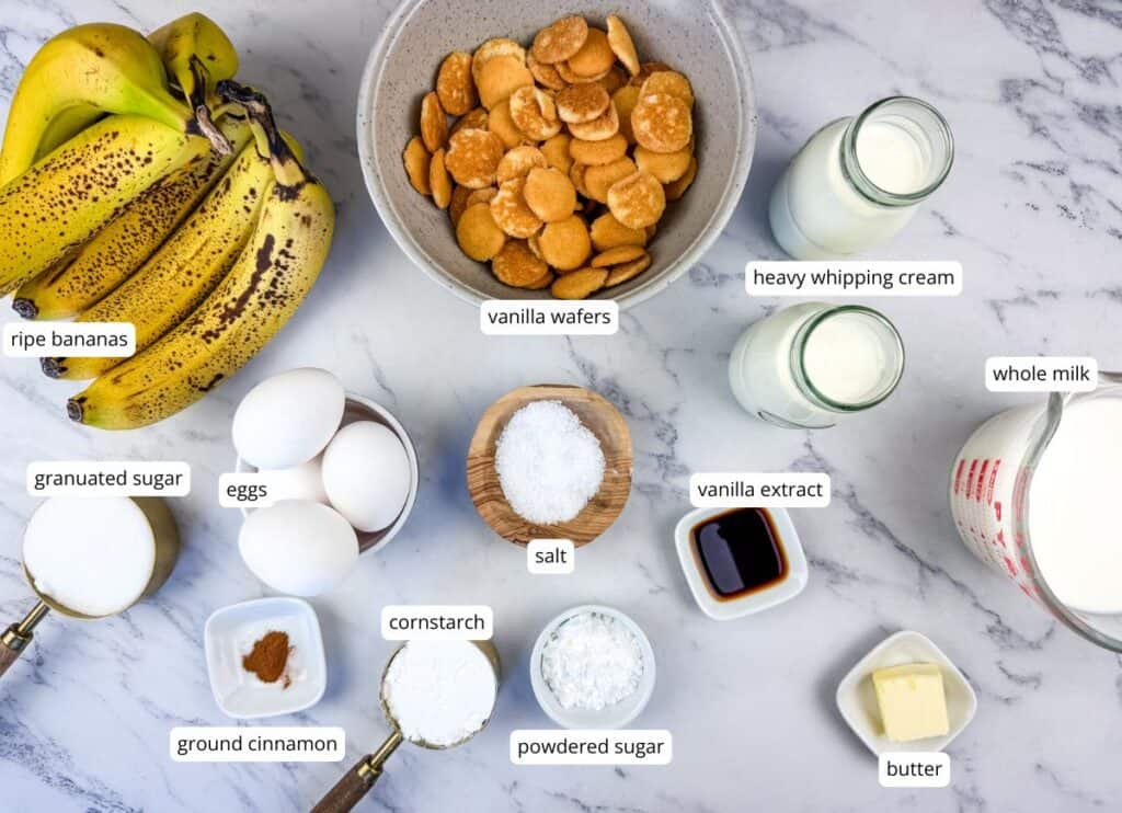 Various ingredients for banana pudding are arranged on a marble surface, including bananas, vanilla wafers, eggs, milk, cream, sugar, cornstarch, cinnamon, salt, vanilla, and butter.
