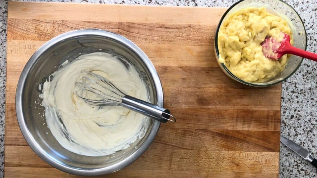 A metal bowl with whisked cream and a whisk sits on a wooden board; next to it is a glass bowl with yellow dough and a red spatula.