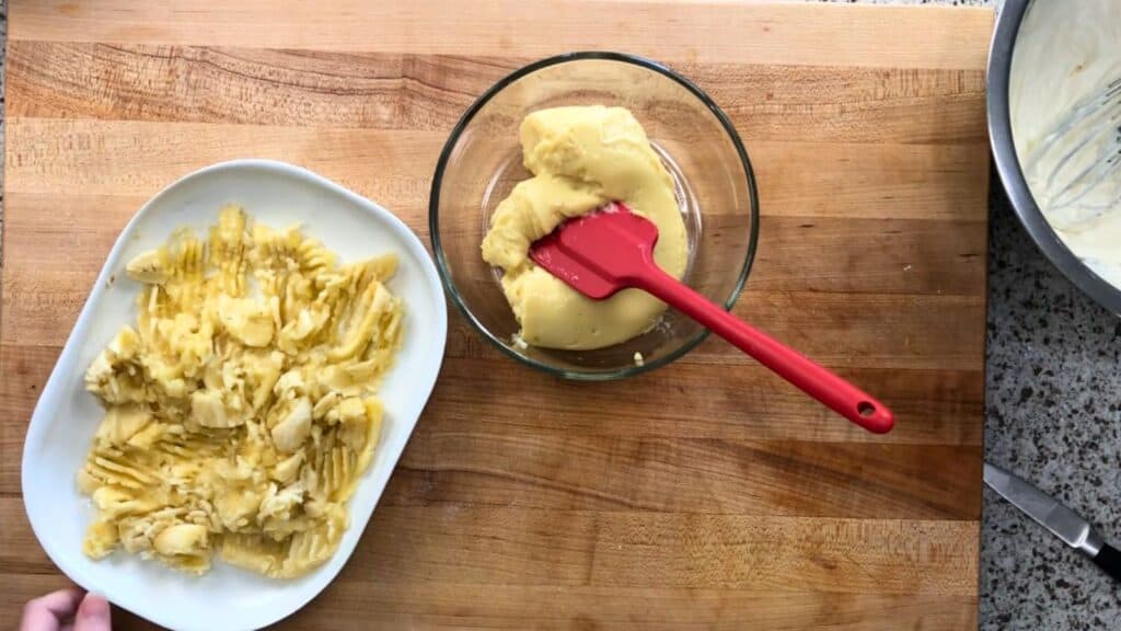 A bowl of mashed potatoes next to a glass bowl of butter with a red spatula, placed on a wooden cutting board.