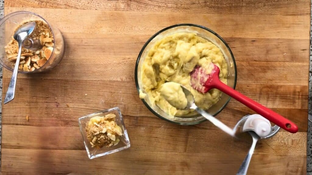 A bowl of banana pudding being prepared with a red spatula, surrounded by crushed cookies in a glass dish and a spoon on a wooden surface.