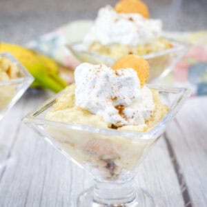 Individual glass cups filled with banana pudding, topped with whipped cream, a vanilla wafer, and a sprinkle of cinnamon, placed on a white wooden surface.