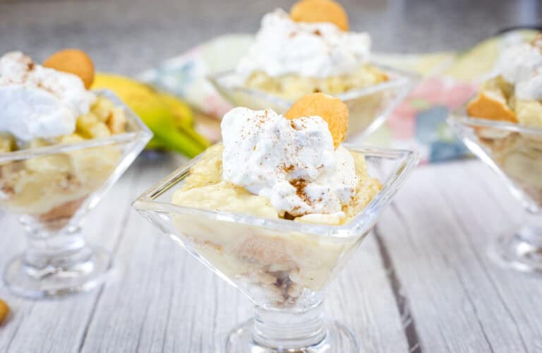 Individual glass cups filled with banana pudding, topped with whipped cream, a vanilla wafer, and a sprinkle of cinnamon, placed on a white wooden surface.