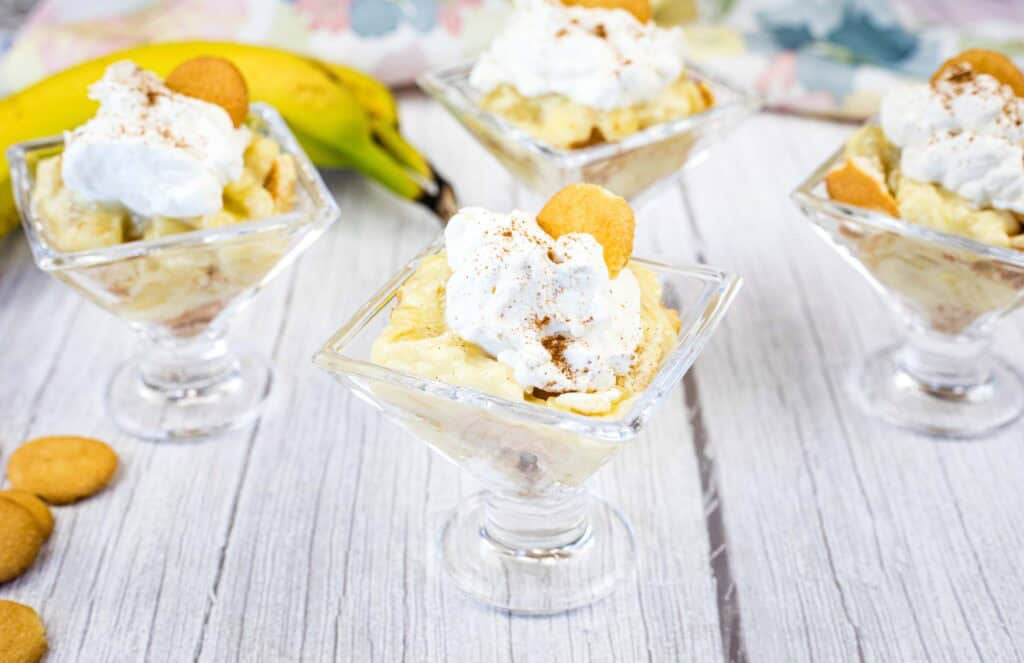 Four glass dessert cups filled with banana pudding, topped with whipped cream, a vanilla wafer, and a sprinkle of cinnamon, are arranged on a white wooden surface.