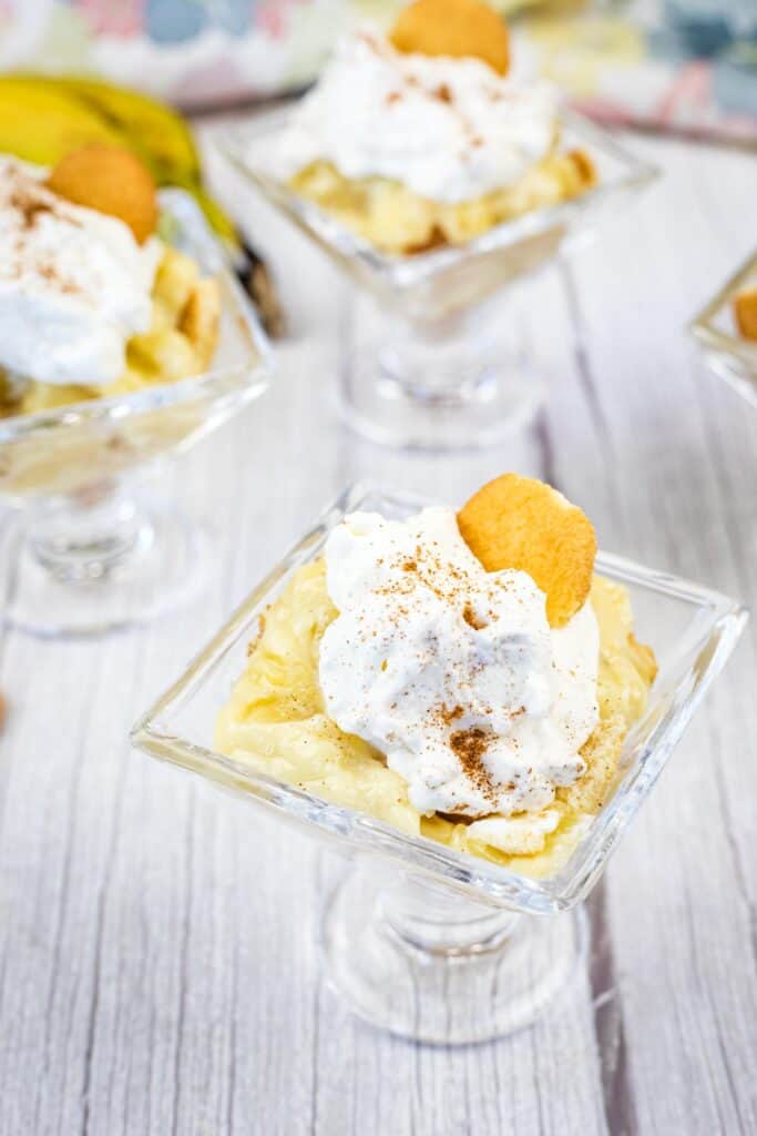 Individual servings of banana pudding topped with whipped cream and a vanilla wafer, presented in small glass dishes on a light wooden surface.