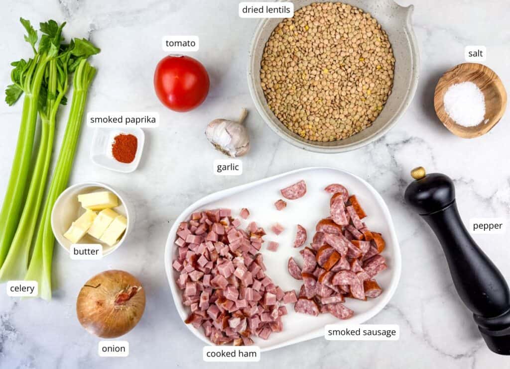 A marble surface displays labeled ingredients for a lentil dish, including dried lentils, tomato, celery, onion, garlic, butter, smoked paprika, cooked ham, smoked sausage, salt, and pepper.