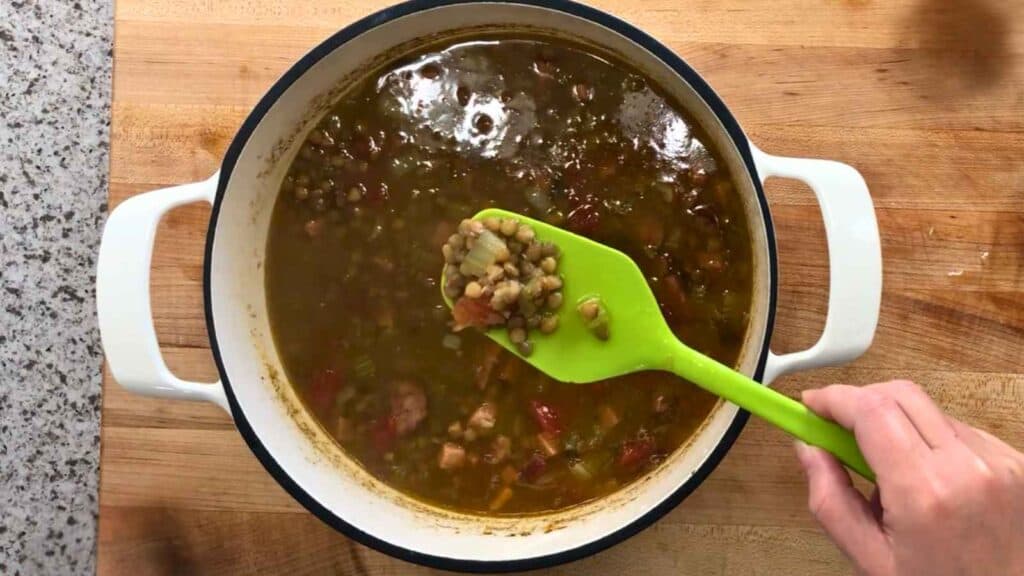 A hand holds a green spoon with lentil soup above a white pot containing more soup, set on a wooden surface.