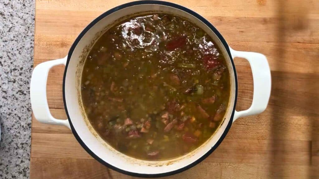 A white pot filled with cooked lentil soup, with visible pieces of vegetables and meat, sits on a wooden countertop.