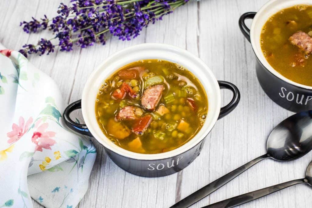 A bowl of vegetable and sausage soup on a light wooden table, with a floral napkin, lavender, and black spoons nearby.