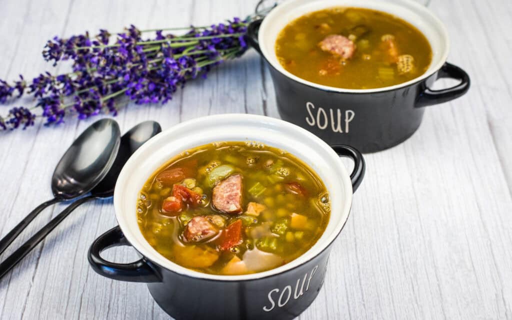 Two black bowls labeled “soup” filled with chunky vegetable and sausage soup, placed on a light wooden surface beside three spoons and a bunch of purple flowers.