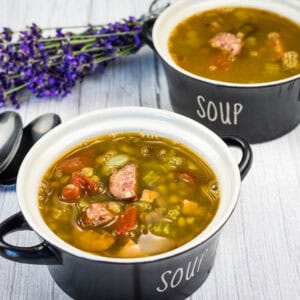 Two black bowls labeled “soup” filled with chunky vegetable and sausage soup, placed on a light wooden surface beside three spoons and a bunch of purple flowers.