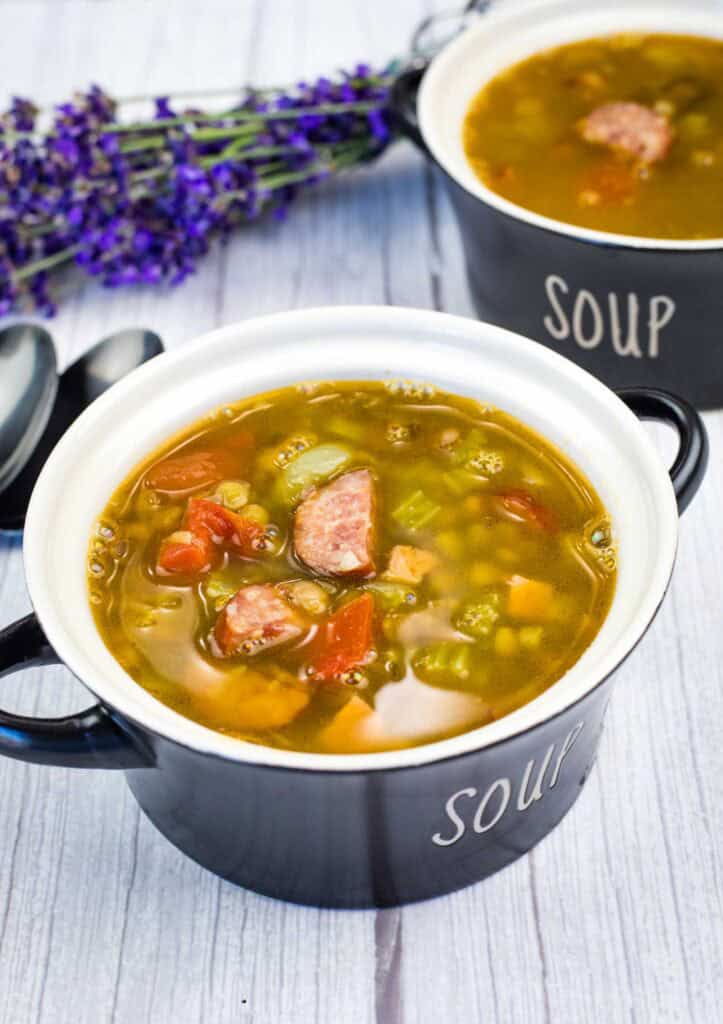 A black bowl labeled “soup” filled with vegetable and sausage soup sits on a light wooden table, with another bowl and purple flowers in the background.
