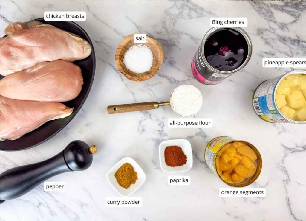 Raw chicken breasts, canned Bing cherries, pineapple, orange segments, salt, flour, black pepper, curry powder, and paprika arranged on a marble surface.