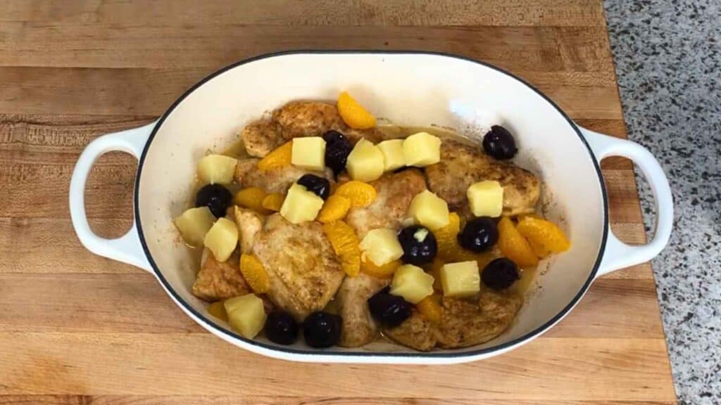 A white baking dish contains cooked chicken pieces topped with pineapple chunks, orange segments, and dark cherries, placed on a wooden countertop next to a speckled surface.