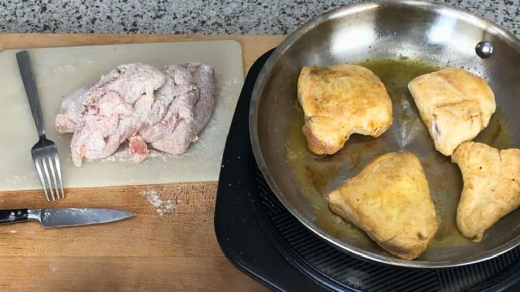 Three pieces of chicken are browning in a pan; floured raw chicken and a fork are on a cutting board nearby, with a knife resting on the countertop.