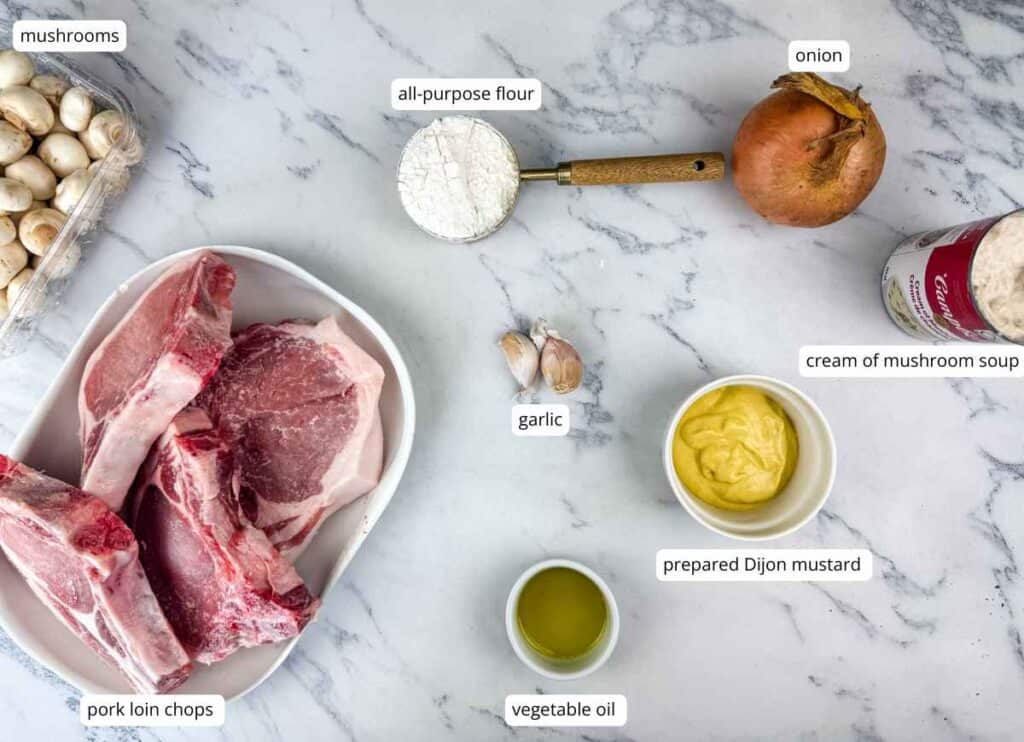 Raw pork loin chops, mushrooms, an onion, all-purpose flour, garlic cloves, vegetable oil, Dijon mustard, and a can of cream of mushroom soup arranged on a white marble surface.