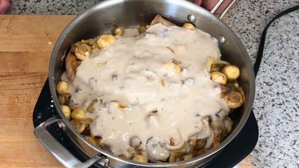 A stainless steel pan on a stovetop contains mushrooms, onions, and meat, topped with a creamy white sauce.