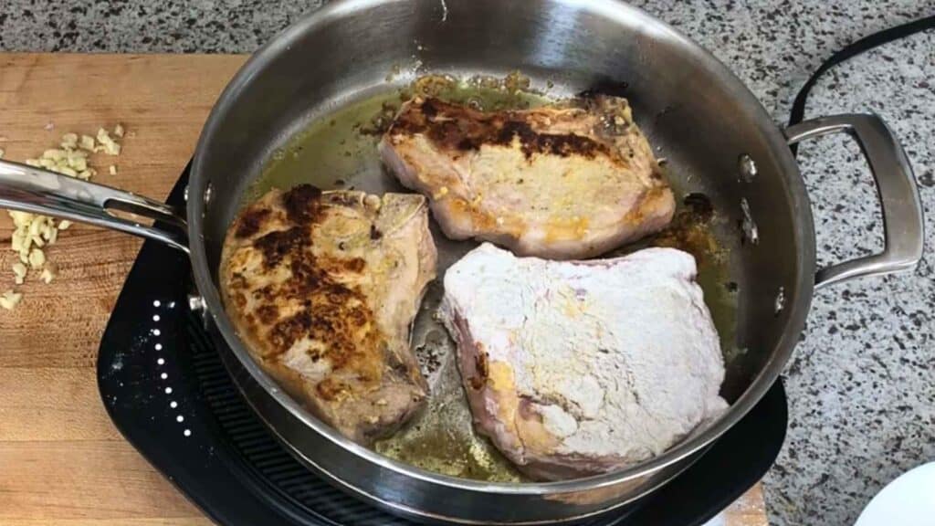 Three pork chops, one coated in flour, are cooking in a stainless steel pan on a stovetop, with a cutting board and chopped garlic visible nearby.