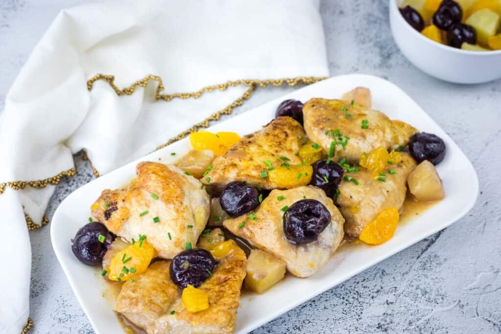 Plated chicken thighs garnished with cherries, orange segments, and chopped herbs on a white dish, with a small bowl of fruit and a white napkin in the background.