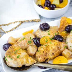 Plated chicken pieces garnished with fresh herbs, orange slices, cherries, and pineapple chunks, with a serving spoon; a bowl of fruit sits in the background.