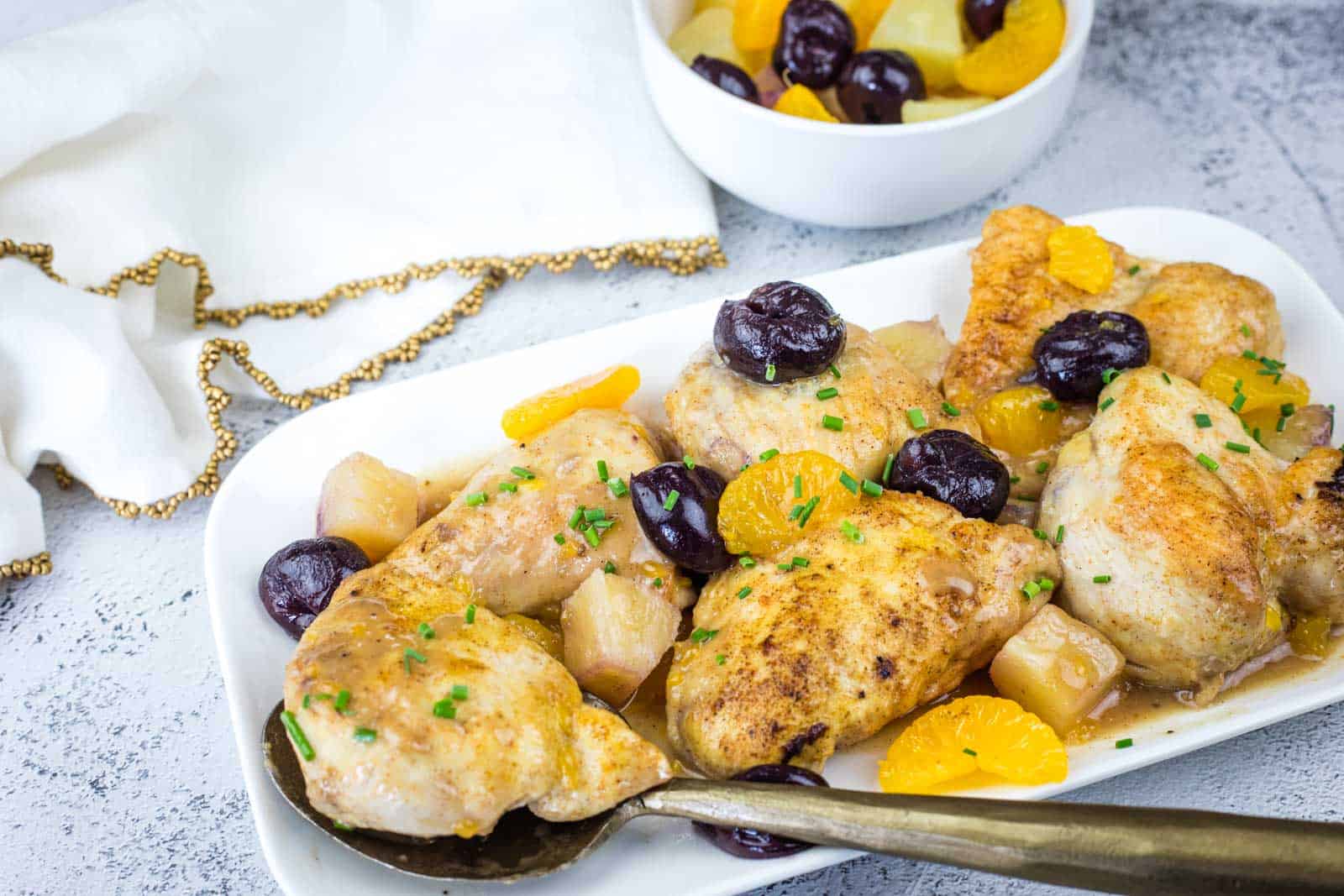 Plated chicken pieces garnished with fresh herbs, orange slices, cherries, and pineapple chunks, with a serving spoon; a bowl of fruit sits in the background.