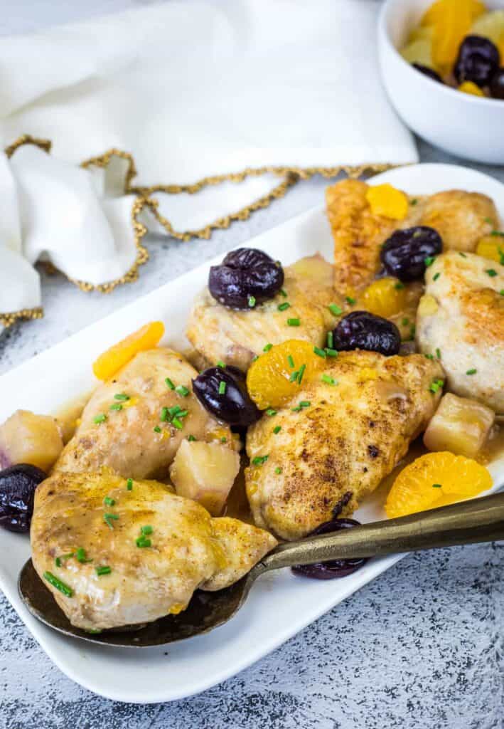Cooked chicken breasts garnished with canned mandarin oranges, dark cherries, and chives on a white platter, with a serving spoon and a bowl of fruit in the background.