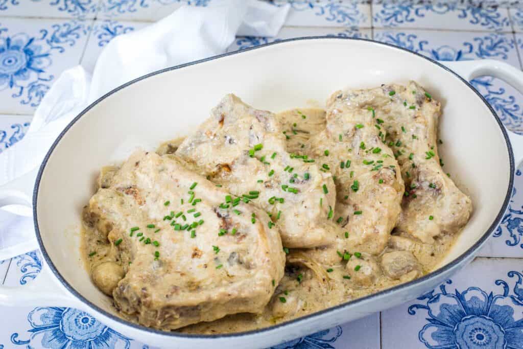 Pork chops in a creamy white sauce, garnished with chopped chives, served in a white oval dish on a blue-patterned tiled surface.
