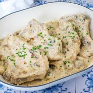 Oval dish with pieces of pork in a creamy sauce, garnished with chopped chives, placed on a blue and white patterned surface.