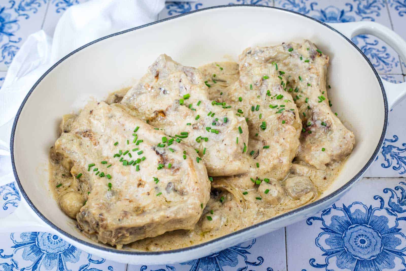 Oval dish with pieces of pork in a creamy sauce, garnished with chopped chives, placed on a blue and white patterned surface.