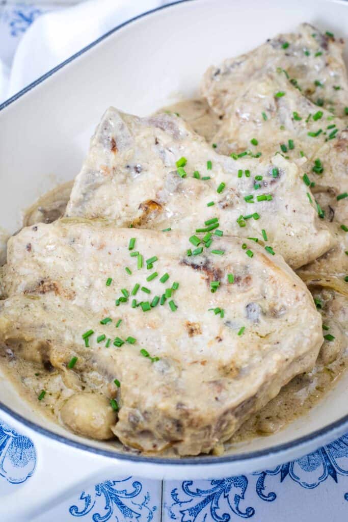 Pork chops covered in creamy mushroom sauce, garnished with chopped chives, served in a white dish.
