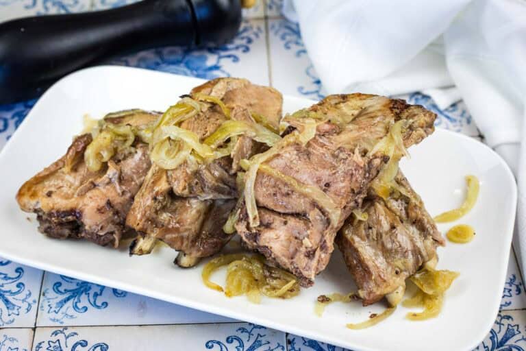 Three pieces of cooked meat with sautéed onions are arranged on a white rectangular plate, placed on a blue and white patterned surface.